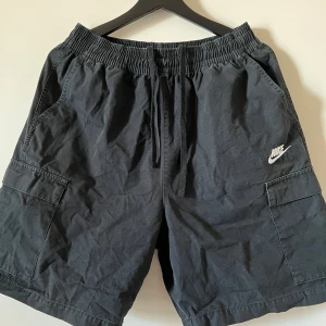 Svarta shorts från Nike - Säljer ett par svarta shorts från Nike med elastisk midja och dragsko. Shortsen har praktiska fickor på sidorna och en bakficka. Perfekta för en avslappnad stil.