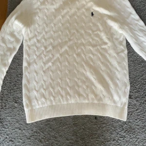Vit stickad tröja från Polo Ralph Lauren - Säljer en stilren vit stickad tröja från Polo Ralph Lauren med det klassiska broderade logotypen på bröstet. Tröjan har en ribbad kant vid ärmslut och nederkant, perfekt för en elegant look. Passar utmärkt för kyligare dagar.