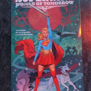 Supergirl: Woman of Tomorrow - Säljer serieboken 'Supergirl: Woman of Tomorrow' av Tom King, med illustrationer av Bilquis Evely och Matheus Lopes. Omslaget visar Supergirl i en dynamisk pose med en röd mantel och en stor röd planet i bakgrunden. Perfekt för fans av superhjälteberättelser och fantastiska illustrationer.