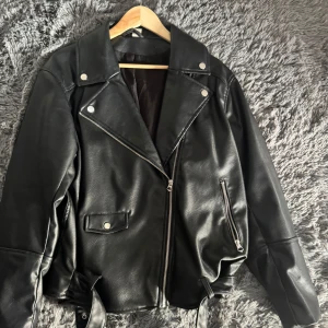 Svart skinnjacka med dragkedja - Snygg svart skinnjacka med dragkedja och knappar. Jackan har en klassisk bikerstil med långa ärmar och flera fickor. 