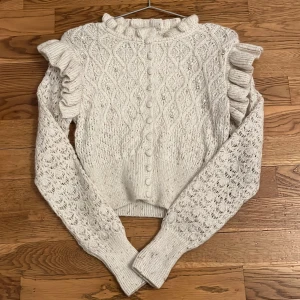 Vit stickad tröja från Zara - Säljer en vit stickad tröja med guld trådar från Zara med vackra volangdetaljer på axlarna och pärlor på framsidan. Tröjan har långa ärmar med ett fint mönster och en volang krage. Perfekt för att ge en elegant touch till din outfit.