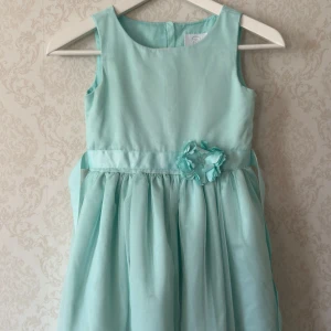 Mintgrön ärmlös klänning med blomdetalj - Söt mintgrön klänning i polyester med en vacker blomdetalj i midjan. Klänningen är ärmlös och har en luftig kjol som ger en fin volym. Perfekt för speciella tillfällen!