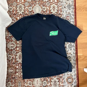 Mörkblå t-shirt från Stussy - Säljer en mörkblå t-shirt från Stussy med en grön logga på bröstet och en stor grön krossad burk på ryggen. Perfekt för en avslappnad stil. T-shirten är kortärmad och gjord av bomull. Tröjan är nästan helt ny!