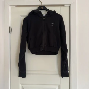 Svart croppad hoodie från Gymshark - Säljer en svart croppad hoodie från Gymshark. Den har en dragkedja framtill och en praktisk huva. Perfekt för en sportig look eller avslappnade dagar. Långärmad design med ribbade muddar.