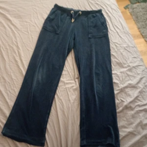 Mörkblå mjukisbyxor från Juicy Couture - Snygga mörkblå mjukisbyxor från Juicy Couture med breda ben och praktiska fickor både fram och bak. Byxorna har en bekväm dragsko i midjan för justerbar passform. Perfekta för en avslappnad stil. Har hål brevid ena bakfickan och är lite slitna vid benen.