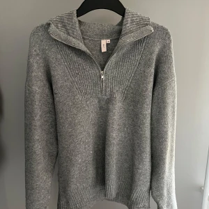 Grå stickad tröja från Nelly - Säljer en stilren grå stickad tröja från Nelly med dragkedja och ribbade detaljer. Perfekt för kyligare dagar med sin mjuka och bekväma passform. Tröjan har långa ärmar och en hög krage som kan bäras uppvikt eller nedvikt.