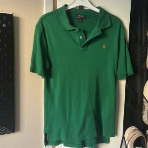 Grön pikétröja från Ralph Lauren - Säljer en klassisk grön pikétröja från Ralph Lauren. Tröjan har korta ärmar och en liten broderad logotyp i orange på bröstet. Perfekt för en stilren och avslappnad look. Hör av dig vid intresse!