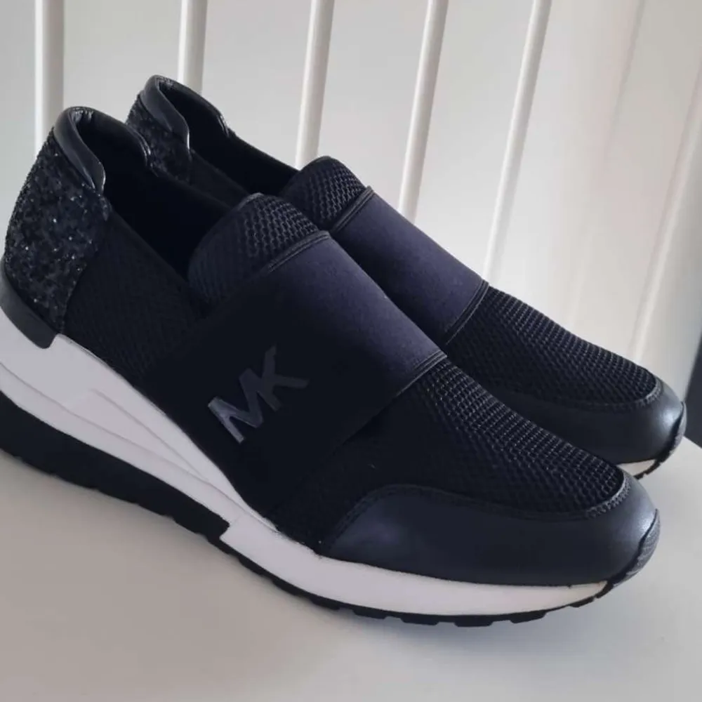 Snygga svarta slip-ins från Michael Kors med glittrig detalj på hälen och elastiskt band med MK-logga över foten. Skorna har en vit och svart sula som ger en sportig look. NYPRIS 2200. Kengät.