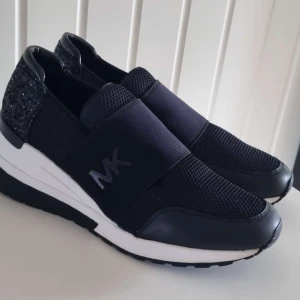 Svarta skor från Michael Kors - Snygga svarta slip-ins från Michael Kors med glittrig detalj på hälen och elastiskt band med MK-logga över foten. Skorna har en vit och svart sula som ger en sportig look. NYPRIS 2200