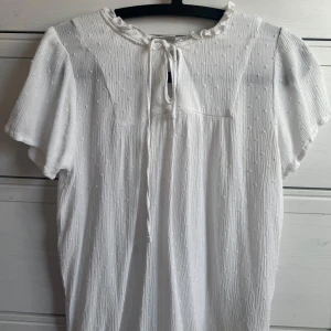 Vit blus från JDY - Säljer en söt vit blus från JDY med korta ärmar och knytning i nacken. Blusen har ett diskret prickigt mönster och en liten volangdetalj vid halsen. Perfekt för en somrig look! 🌼