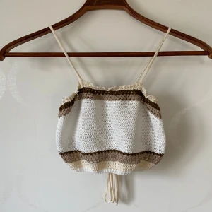Randig virkad linnetopp - Säljer en söt virkad linnetopp med smala axelband. Toppen har ett randigt mönster i vitt, beige och brunt och är försedd med snörning i ryggen för en justerbar passform. Perfekt för varma sommardagar!