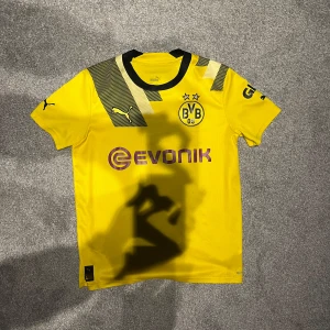 Borussia Dortmund fotbollströja storlek 164 från Puma  - Säljer en gul Borussia Dortmund fotbollströja från Puma med svart tryck och klubbmärke. Tröjan har korta ärmar och är tillverkad i ett lätt och andningsbart material. Perfekt för fotbollsfans som vill visa sitt stöd för laget. 🖤💛