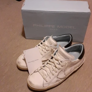 Vita sneakers från Philippe Model strl 44 - Snygga vita sneakers från Philippe Model med svarta detaljer på hälen. Skorna har klassisk snörning och en stilren design som passar perfekt till vardags. De är tillverkade i skinn och har en bekväm passform. Slitna bak på sulorna, bild, men det märks inte vid användning. Originallåda finns. 