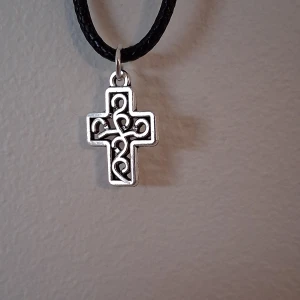 Silverfärgat korshalsband - Snyggt halsband med ett silverfärgat korshänge. Korset har ett intrikat mönster och hänger på en svart snodd. Perfekt för att ge en stilren touch till din outfit.