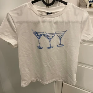 Vit t-shirt med martiniglas från Shein - Säljer en vit t-shirt från Shein med ett coolt tryck av tre blå martiniglas på framsidan. Perfekt för en avslappnad stil med en touch av festkänsla. T-shirten är kortärmad och gjord i bomull.