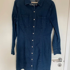 Blå jeansklänning med knappar - Snygg blå jeansklänning med långa ärmar och knappar framtill. Slimfit med stretch. Detaljbild något missvisande i färg - den är mer mörkblå likt helbilderna