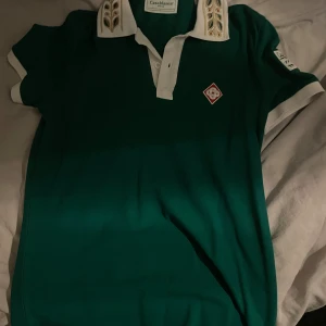 Grön pikétröja från Casablanca - Säljer en snygg grön pikétröja från Casablanca med vita detaljer och ett unikt mönster på kragen. Tröjan har korta ärmar och en liten logotyp på bröstet. Perfekt för en stilren look! Den har aldrig använts!