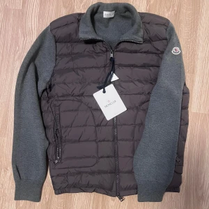 Grå cardigan Moncler - Snygg grå cardigan från Moncler med dragkedja och ribbade ärmslut. Jackan har en kombination av quiltat material och stickade ärmar, vilket ger en unik look. Perfekt för kyligare dagar.Det är i väldigt bra skick det är storlek S. Såklart funkar det att skanna moncler märket