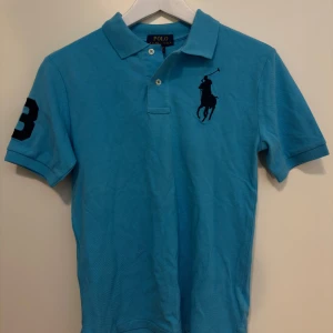 Blå pikétröja från Ralph Lauren - Säljer en snygg blå pikétröja från Ralph Lauren med korta ärmar och klassisk krage. Tröjan har en broderad logga på bröstet och en stor siffra 3 på ärmen. Perfekt för en stilren look!