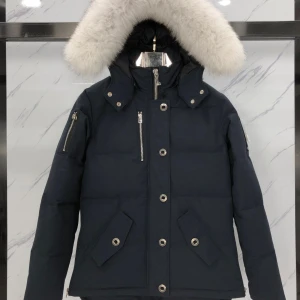 Svart dunjacka med pälsdetalj - Snygg svart dunjacka från Moose Knuckles med en lyxig vit pälsdetalj på huvan. Jackan har dragkedja och knappar framtill samt praktiska fickor med knappar. Perfekt för kalla dagar med sin quiltade design och långa ärmar.