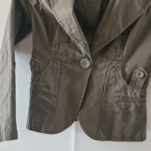 Olivgrön kavaj med knappar - Snygg olivgrön kavaj med stora knappar framtill och på ärmarna. Den har en klassisk krage och två fickor med lock. Perfekt för en stilren look.