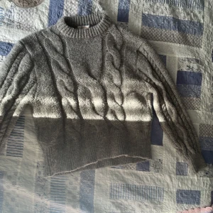 Grå stickad tröja med flätmönster - Säljer en mysig grå stickad tröja med ett snyggt flätmönster på framsidan. Tröjan har långa ärmar och en ribbad krage samt ribbade muddar vid ärmslut och nederkant. Perfekt för kyliga dagar!