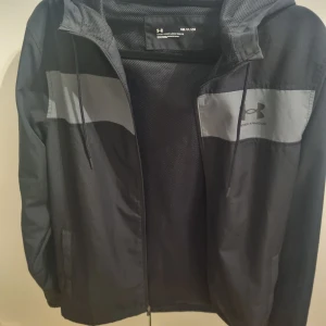 Svart herrjacka från Under Armour - Svart jacka från Under Armour med grå detaljer och huva. Jackan har dragkedja och snöre vid luvan.