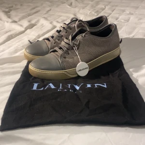 Grå sneakers från Lanvin - Snygga grå sneakers från Lanvin med en unik textur och snörning. Skorna har en ljusgummisula och en grå tåhätta som ger en stilren look. Perfekta för en casual stil med en touch av lyx. Dustbag tillkommer vid köp. Storlek 40 men sitter som 41. Bara hör av er vid frågor o funderingar!