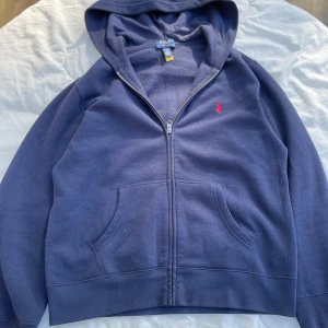 Mörkblå zip från Ralph Lauren - Säljer en snygg mörkblå hoodie från Ralph Lauren med dragkedja och huva. Den har en röd broderad logga på bröstet och praktiska fickor framtill. Perfekt för en avslappnad stil. Tröjan är i perfekt skick och inga skador. Skriv till mig om fler bilder eller frågor💛