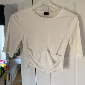 Croppad top från Gina  - Säljer en stilren vit ribbad topp från Gina Tricot. Toppen har trekvartsärmar och en snygg omlott-detalj framtill som ger en modern touch. Perfekt för att matcha med både jeans och kjol.