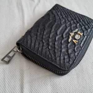 Svart plånbok med dragkedja - zadig Voltaire  - Snygg svart plånbok med dragkedja och textur som liknar reptilmönster. Den har en metallisk ZV logotyp på framsidan och flera fack inuti för kort och kontanter. Perfekt för att hålla dina saker organiserade och säkra.