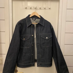 Mörkblå jeansjacka från Arket - Mörkblå jeansjacka med klassisk design. Jackan har knappar framtill och två bröstfickor med knappstängning.