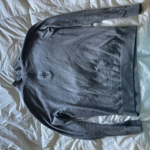 Grå tröja från Tommy Hilfiger - Säljer en stilren grå tröja från Tommy Hilfiger i bomull och silke. Tröjan har en halv dragkedja vid halsen och långa ärmar. Perfekt för en avslappnad och snygg look.