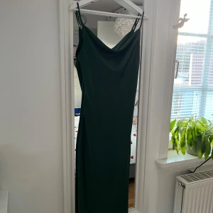 Mörkgrön klänning - Elegant mörkgrön klänning med smala axelband och en stilren design. Perfekt för en kväll ute eller en speciell tillställning. Klänningen har en vacker drapering och ger en sofistikerad look.