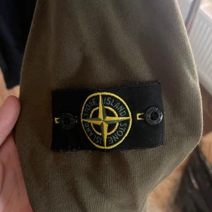 Olivgrön tröja från Stone Island - Säljer en snygg olivgrön tröja från Stone Island med det ikoniska märket på ärmen. Tröjan har en rund halsringning och långa ärmar. Perfekt för en stilren och avslappnad look.