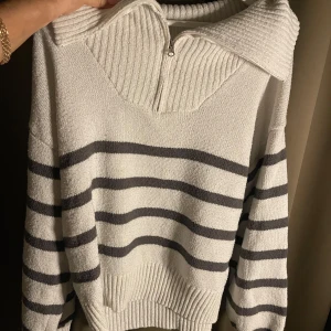 Vit och grå randig tröja med dragkedja - Säljer en snygg vit och grå randig tröja med dragkedja vid halsen. Tröjan har en ribbad krage och långa ärmar, perfekt för en stilren look. Passar bra till både jeans och kjol.