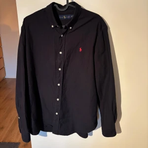 Skjorta från Ralph Lauren - Snygg svart skjorta från Ralph Lauren med klassisk krage och knappar framtill. Den har en broderad logotyp i rött på bröstet och långa ärmar. Perfekt för en stilren look. Skjortan är mörkblå men den ser svart ut på bilderna