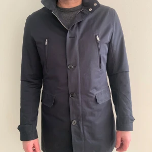 Svart rock från J.Lindeberg - Snygg svart rock från J.Lindeberg med knappar och dragkedja framtill. Rocken har en stilren design med långa ärmar och två fickor med dragkedja på bröstet. Skriv om ni vill ha fler bilder!!