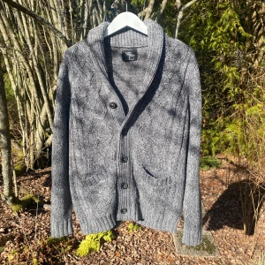 Grå kabelstickad kofta från Cedar Wood State - Säljer en snygg grå kabelstickad kofta från Cedar Wood State. Den har en ribbad krage och knappar framtill. Perfekt för kyliga dagar med sin stickade design och praktiska fickor. Passar bra till både jeans och chinos.