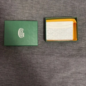 Vit plånbok  - Säljer en elegant vit plånbok från G🅾️yard med deras ikoniska mönster. Plånboken kommer i en snygg grön förpackning och inkluderar ett äkthetskort. Perfekt för att hålla dina kort och kontanter organiserade.