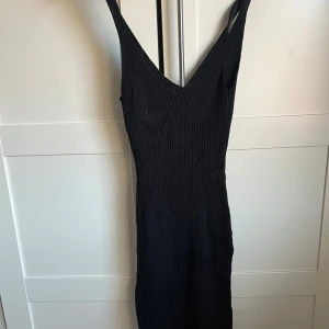 Svart ribbad klänning - Snygg svart ribbad klänning med smala axelband och v-ringad design. Perfekt för en stilren look. Klänningen har en tight passform som framhäver figuren.