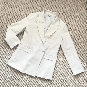 Vit kavaj med knappar - Snygg vit kavaj med klassisk design och två knappar framtill. Den har långa ärmar och två fickor med lock. Perfekt för en stilren look.