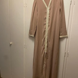 Beige långklänning kaftan abaya - Elegant beige långklänning med v-ringad design och dekorativ kant i guld. Klänningen har långa ärmar och ett stilrent fall, perfekt för speciella tillfällen. Den är använd ungefär två gånger men har inga tecken på användning.