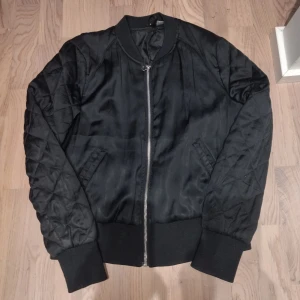 Svart bomberjacka från Divided - Snygg svart bomberjacka från Divided med quiltade ärmar och dragkedja framtill. Jackan har en klassisk bomberstil med ribbade muddar vid ärmslut och nederkant. Perfekt för en cool och avslappnad look.