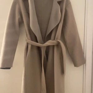 Beige kappa från Nelly - Säljer en stilren beige kappa från Nelly i storlek XS. Den har en elegant krage och ett bälte i midjan för en snygg passform. Perfekt för höst och vår! Endast använd ett par fåtal gånger, mycket bra skick. För att se passformen kolla bild 4 & 5 - exakt samma jacka fast i svart!