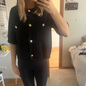 Svart kortärmad topp från Massimo Dutti  med guldknappar - Snygg svart topp från Massimo Dutti med korta ärmar och dekorativa guldknappar. Den har en texturerad yta som ger en unik look. Perfekt för att lyfta vilken outfit som helst.