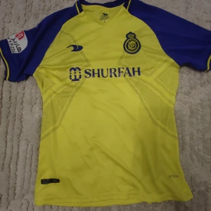 Al Nassr 22/23 Hemma tröja - Säljer en gul och blå Al Nassr fotbollströja från Duneus. Tröjan har korta ärmar och klubbens emblem på bröstet. Namnet 'Ronaldo' och nummer 7 pryder ryggen. Perfekt för fotbollsfans