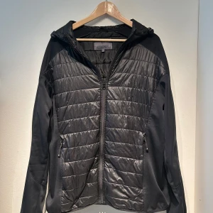 Cardigan - Säljer en svart cardigan från Cross Sportswear i storlek XL. Jackan har en quiltad design med dragkedja och huva. Perfekt för kyligare dagar med sin stilrena look och praktiska fickor.