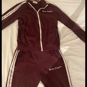 Vinröd äkta tracksuit  från Palm Angels  - Säljer en snygg vinröd träningsjacka från Palm Angels med dragkedja och vita ränder längs ärmarna. Jackan har en normal passform och är perfekt för en sportig look. Den har även Palm Angels-loggan tryckt på bröstet. 