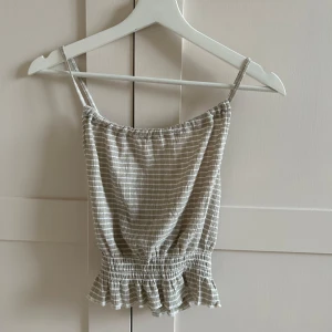 Beige randig linnetopp från Gina Tricot - Säljer en söt beige randig linnetopp från Gina Tricot. Toppen har smala axelband och en volangdetalj vid nederkanten. Jätteskönt bas linne!!